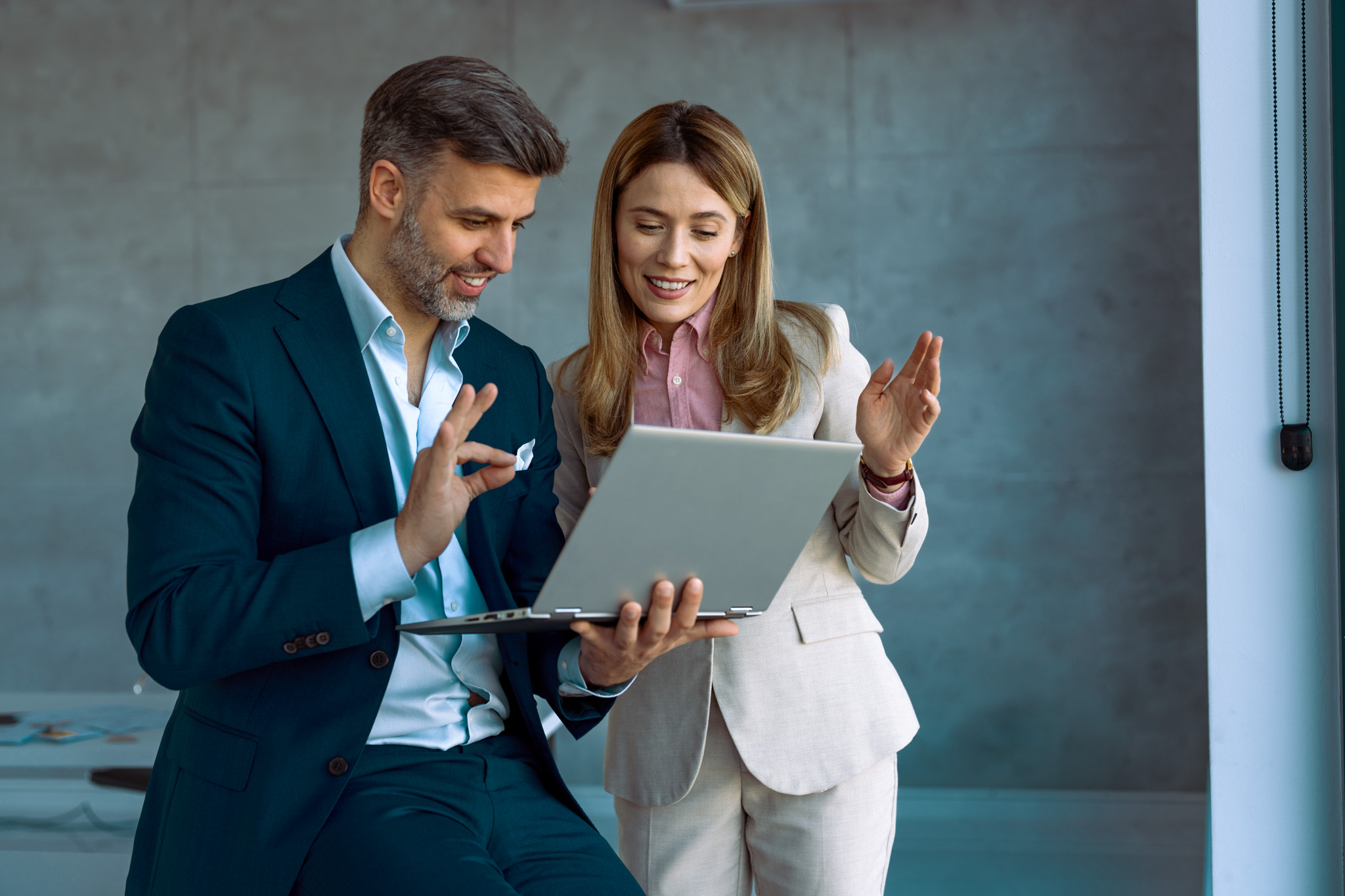 A mid adult Caucasian businessman and businesswoman engage in a discussion over a laptop, dressed in formal attire, symbolizing teamwork in a modern office environment.